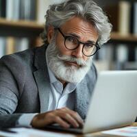An elderly man focuses on his laptop while sitting in a stylish office filled with books photo