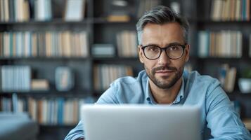 Man with graying hair wearing glasses looks directly at camera while working on laptop in an office photo