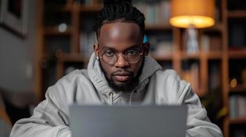 A man with dreadlocks wearing glasses sits at a desk in front of a laptop, looking intently at the screen photo