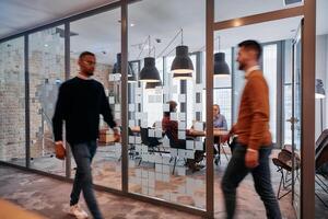 In the dynamic hustle and bustle of a business environment, a group of young business professionals walk down the corridor next to their office, while their colleagues collaborate inside, symbolizing photo