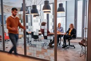 In the dynamic hustle and bustle of a business environment, a group of young business professionals walk down the corridor next to their office, while their colleagues collaborate inside, symbolizing photo