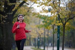 Young beautiful woman running in autumn park and listening to music with headphones on smartphone photo