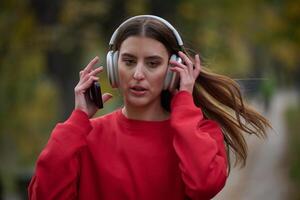 Young beautiful woman running in autumn park and listening to music with headphones on smartphone photo