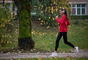 Young beautiful woman running in autumn park and listening to music with headphones on smartphone photo