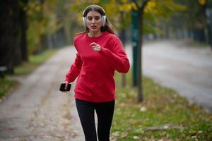 Young beautiful woman running in autumn park and listening to music with headphones on smartphone photo