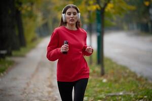 Young beautiful woman running in autumn park and listening to music with headphones on smartphone photo