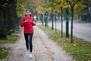 Young beautiful woman running in autumn park and listening to music with headphones on smartphone photo