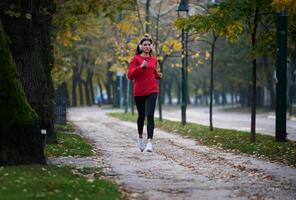 Young beautiful woman running in autumn park and listening to music with headphones on smartphone photo