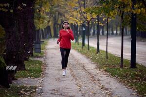 Young beautiful woman running in autumn park and listening to music with headphones on smartphone photo