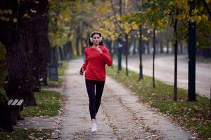 Young beautiful woman running in autumn park and listening to music with headphones on smartphone photo