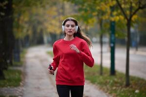 Young beautiful woman running in autumn park and listening to music with headphones on smartphone photo