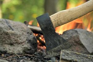 Ax lodged in a log of wood, positioned across from a campfire photo