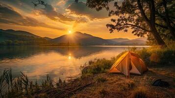 Idyllic camping scene with tent near calm lake during golden hour, sunset behind mountains, sky with warm golden hues, serene and peaceful atmosphere. Camping taken during golden hour photo