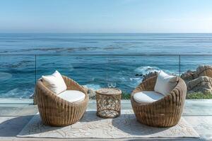 Two round wicker chairs with cushions and a small wicker table with a plant, all set on a wooden deck overlooking a stunning ocean view. photo