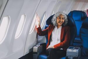 Portrait of a successful Asian businesswoman or entrepreneur in a formal suit on an airplane sitting in business class using a phone, computer laptop. Travel in style, work with grace. photo