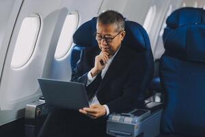 Attractive Asian male passenger of airplane sitting in comfortable seat while working laptop and tablet with mock up area using wireless connection. Travel in style, work with grace. photo