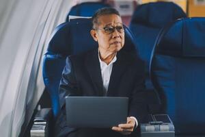 Attractive Asian male passenger of airplane sitting in comfortable seat while working laptop and tablet with mock up area using wireless connection. Travel in style, work with grace. photo