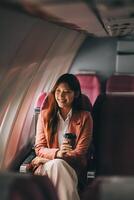 Portrait of a successful Asian businesswoman or entrepreneur in a formal suit on an airplane sitting in business class using a phone, computer laptop. Travel in style, work with grace. photo