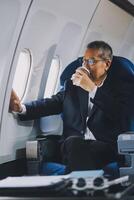 Attractive Asian male passenger of airplane sitting in comfortable seat while working laptop and tablet with mock up area using wireless connection. Travel in style, work with grace. photo