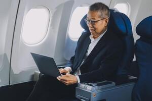 Attractive Asian male passenger of airplane sitting in comfortable seat while working laptop and tablet with mock up area using wireless connection. Travel in style, work with grace. photo