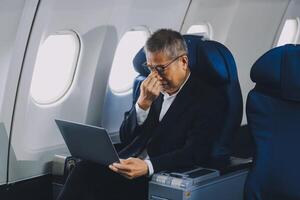 Attractive Asian male passenger of airplane sitting in comfortable seat while working laptop and tablet with mock up area using wireless connection. Travel in style, work with grace. photo
