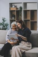 Happy grownup daughter showing content on tablet to mature mother. Two family generations women with digital computer gadget resting on couch together, watching videos, making call photo