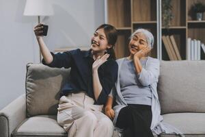Cheerful mother and pretty teenager kid relaxing at cozy home, using mobile phone for Internet communication, resting on sofa with digital gadget, enjoying leisure, looking at camera photo