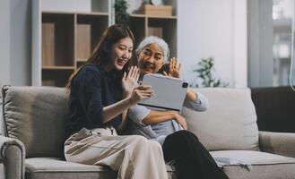 Happy grownup daughter showing content on tablet to mature mother. Two family generations women with digital computer gadget resting on couch together, watching videos, making call photo