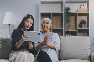 Happy grownup daughter showing content on tablet to mature mother. Two family generations women with digital computer gadget resting on couch together, watching videos, making call photo