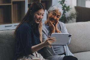 Happy grownup daughter showing content on tablet to mature mother. Two family generations women with digital computer gadget resting on couch together, watching videos, making call photo