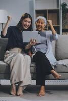 Happy grownup daughter showing content on tablet to mature mother. Two family generations women with digital computer gadget resting on couch together, watching videos, making call photo