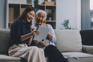 Happy grownup daughter showing content on tablet to mature mother. Two family generations women with digital computer gadget resting on couch together, watching videos, making call photo