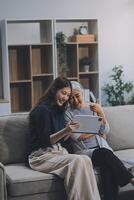 Happy grownup daughter showing content on tablet to mature mother. Two family generations women with digital computer gadget resting on couch together, watching videos, making call photo