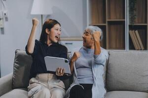 Happy grownup daughter showing content on tablet to mature mother. Two family generations women with digital computer gadget resting on couch together, watching videos, making call photo