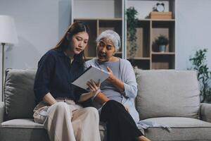 Happy grownup daughter showing content on tablet to mature mother. Two family generations women with digital computer gadget resting on couch together, watching videos, making call photo
