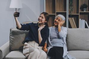 Cheerful mother and pretty teenager kid relaxing at cozy home, using mobile phone for Internet communication, resting on sofa with digital gadget, enjoying leisure, looking at camera photo