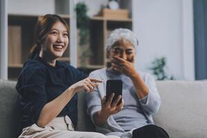 Cheerful mother and pretty teenager kid relaxing at cozy home, using mobile phone for Internet communication, resting on sofa with digital gadget, enjoying leisure, looking at camera photo