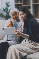 Happy grownup daughter showing content on tablet to mature mother. Two family generations women with digital computer gadget resting on couch together, watching videos, making call photo