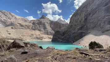 Timelapse view Bolshoi Allo Lake of Fann Mountains in Tajikistan sunny day with clouds. Turquoise high mountain pond surrounded by towering Rocky Cliffs under a vibrant blue sky with scattered clouds video