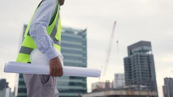 Engineerman in a yellow vest is holding a stack of blueprints while walking on a rooftop. Concept of professionalism and focus, as the man is likely a construction worker or engineer video