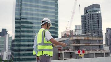 Two Engineer men are standing on a rooftop of a building, one of them is holding a tablet. The other man is wearing a yellow vest. The scene is set in a city with a large building in the background video