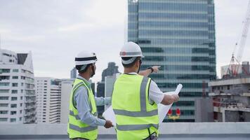 Two engineer men in safety vests are standing on a rooftop looking at blueprints. Scene is serious and focused, as the men are likely discussing plans for a construction project video
