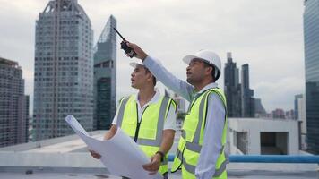 Engineer men are standing on a rooftop, looking at a blueprint. They are wearing safety gear and seem to be discussing the plans. Concept of professionalism and focus video