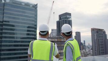 Two engineer men in safety vests are standing on a rooftop of a building. They are looking at something and discussing it video