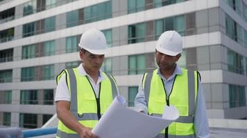 Two engineer men in yellow vests are standing in front of a building, looking at blueprints. Scene is serious and focused video