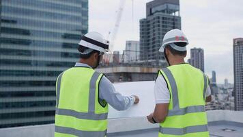 Engineer men in yellow vests are standing on a rooftop, looking at a blueprint. Scene is serious and focused, as the men are likely discussing the details of the building project video