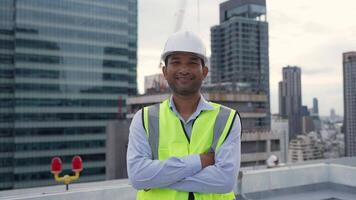 Engineer man in a yellow vest stands on a rooftop in front of a city skyline. He is wearing a hard hat and he is a construction worker. Concept of urban life and the hard work that goes into building video