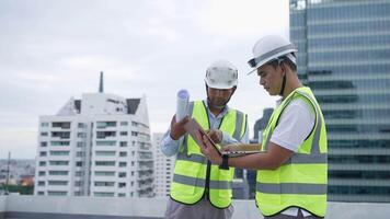Engineer men in yellow vests are looking at a laptop. They are wearing hard hats and are standing on a rooftop video
