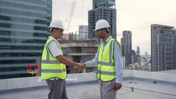 Two engineer men in safety vests walk on a rooftop, one of them holding a clipboard. The scene is set in a city with a large crane in the background. Scene is professional and focused video