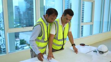 Two engineer men in yellow vests are standing in a large room, looking at blueprints. Scene is serious and focused, as the men are likely discussing plans or making decisions related to their work video
